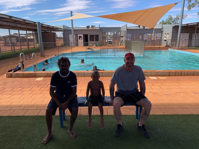 Image: Two men and a little boy sit on the new bench with swimming pool in background and kids swimming.